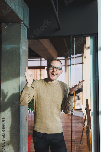 A young man in glasses and casual clothes stands in a modern workspace. Male freelancer works remotely.