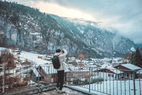 Young tourist man in warm clothes with camera shooting a photo on the background of winter city Lauterbrunnen in Switzerland Europe, Winter landscape, Concept of Travel and holiday