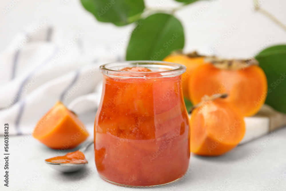 Jar of tasty persimmon jam and ingredients on white table, closeup