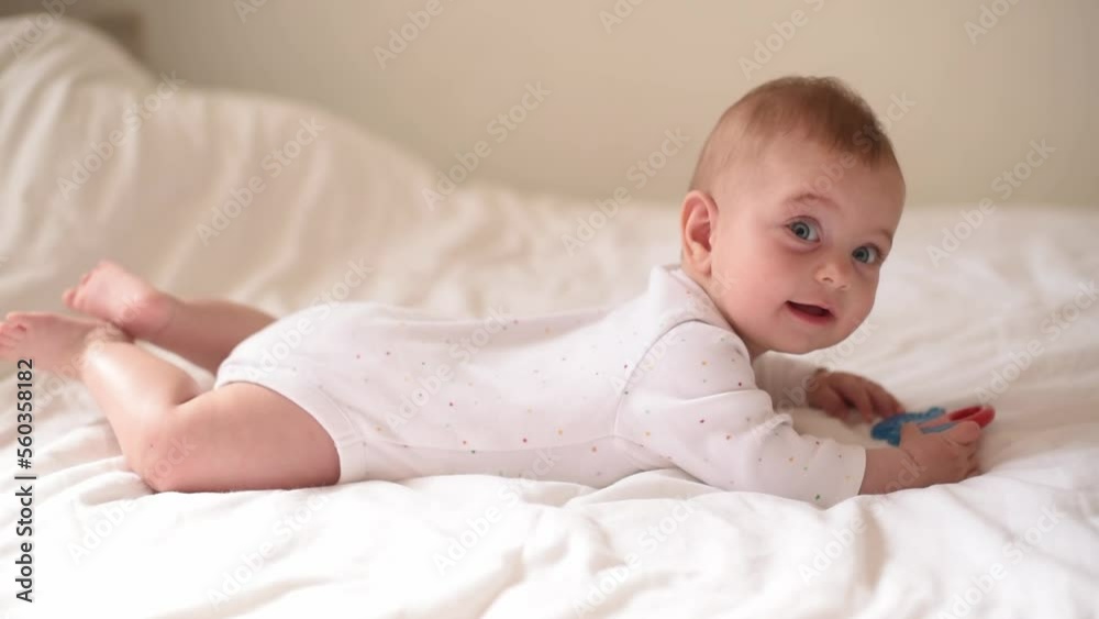 Smiling baby with blue eyes and in white t-shirt lies on belly on bed. Kid takes toy in hand. Close-up. First year of life and knowledge of world around. Side view Smiling baby with blue eyes and in white t-shirt lies on belly on bed. Kid takes toy in hand. Close-up. First year of life and knowledge of world around. Side view