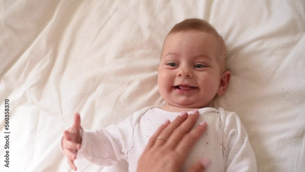 Smiling baby with blue eyes and in white t-shirt lies on bed. Hands of mother caress of kid. Close-up. Love and care about small child. Top view. Smiling baby with blue eyes and in white t-shirt lies on bed. Hands of mother caress of kid. Close-up. Love and care about small child. Top view.