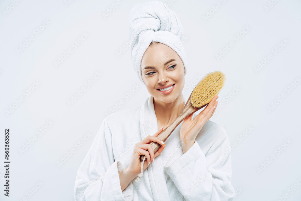 Beautiful glad woman with healthy skin and toothy smile holds wooden massage brush for body, wears bath robe and wrapped soft towel on head, stands against white background. Beauty, care concept Beautiful glad woman with healthy skin and toothy smile holds wooden massage brush for body, wears bath robe and wrapped soft towel on head, stands against white background. Beauty, care concept