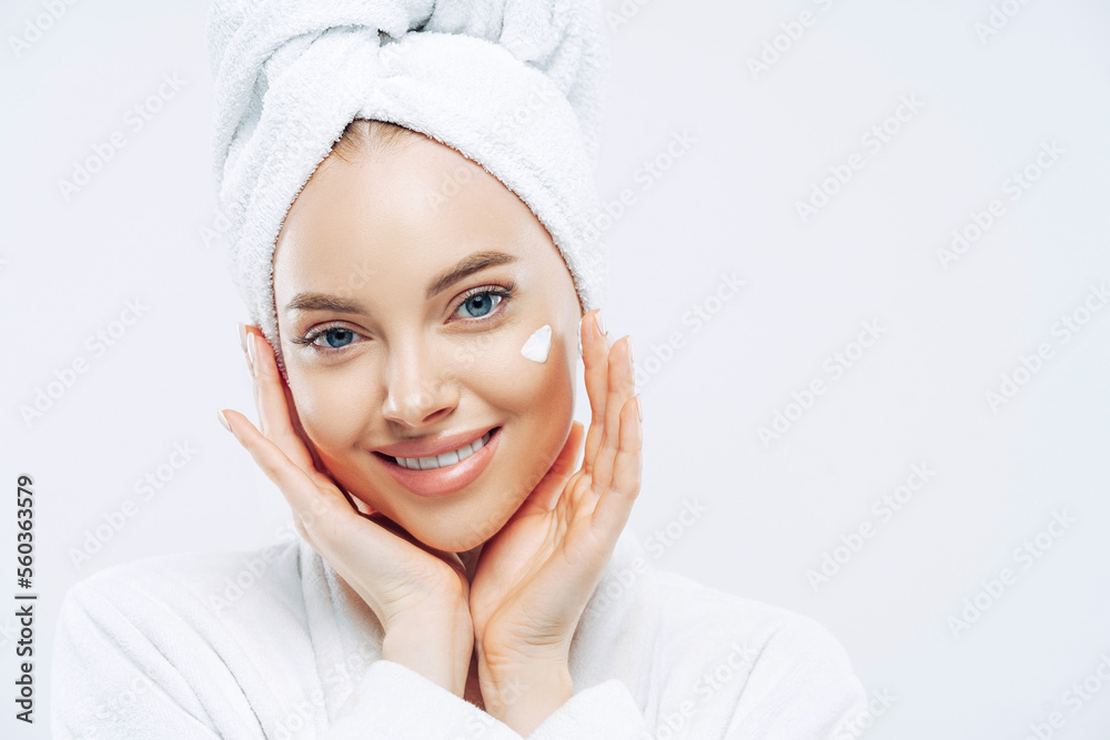 Close up portrait of cheerful female with natural beauty, touches cheeks gently, applies face cream for soft skin, wears bath towel on head, dressed in robe, stands indoor. Beauty treatment, lifestyle Close up portrait of cheerful female with natural beauty, touches cheeks gently, applies face cream for soft skin, wears bath towel on head, dressed in robe, stands indoor. Beauty treatment, lifestyle