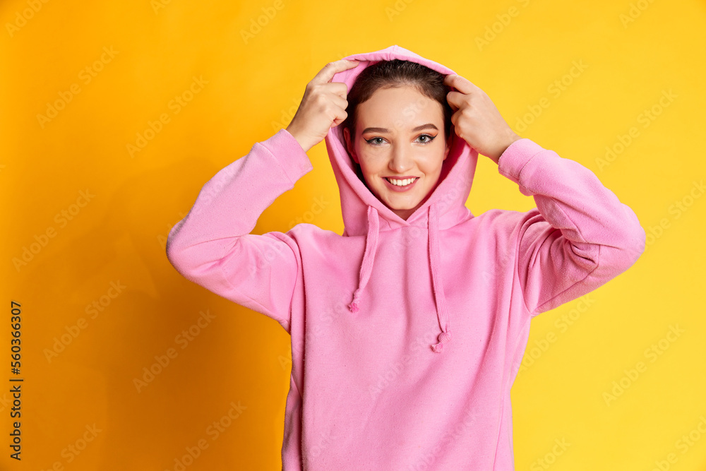 Portrait of young beautiful girl posing in pink hoodie, smiling over yellow studio background. Concept of youth, emotions Portrait of young beautiful girl posing in pink hoodie, smiling over yellow studio background. Concept of youth, emotions