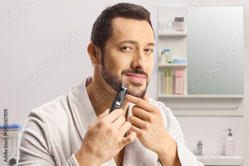 Young man in a bathrobe using a beard trimmer in a bathroom photo