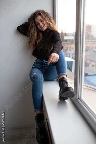 Full length image of a cheerful gorgeous young woman with curly hair, wear black blouse and jeans, posing near windows.