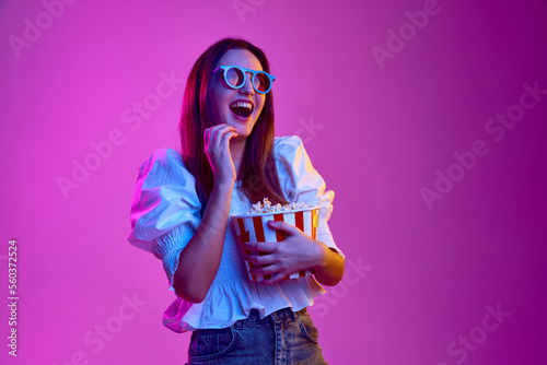 Comedy, laugh. Portrait of young emotive girl posing in 3D glasses with popcorn basket over pink background in neon light. Concept of emotions, youth, lifestyle