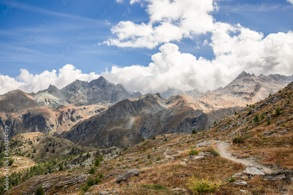 Alpine hiking path on granite slope covered with yellowed withered autumn grass and rare dwarf trees in Aosta Valley, Gran Paradiso National Park, Italy