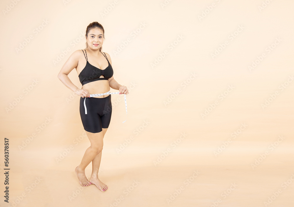 Fototapeta premium Beautiful indian girl measuring her waistline with measure tape isolated on beige background. Weight loss, slim body, healthy lifestyle concept, Full length.