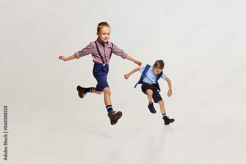 Joyful boys, children in stylish classical clothes playing together, running, jumping over grey studio background. Concept of game, childhood, friendship