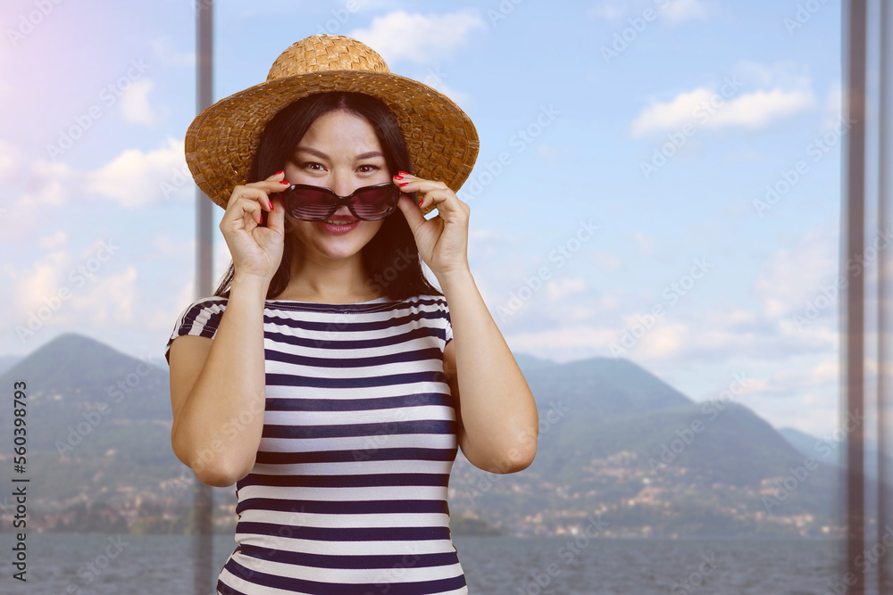 Portrait of a young asian woman in straw hat and striped shirt puts on sunglasses. Mountain landscape background from indoors.