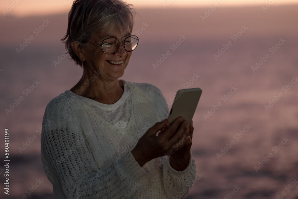 Portrait of cheerful senior woman using phone at sunset standing outdoors at sea - elderly lady reading message on smartphone