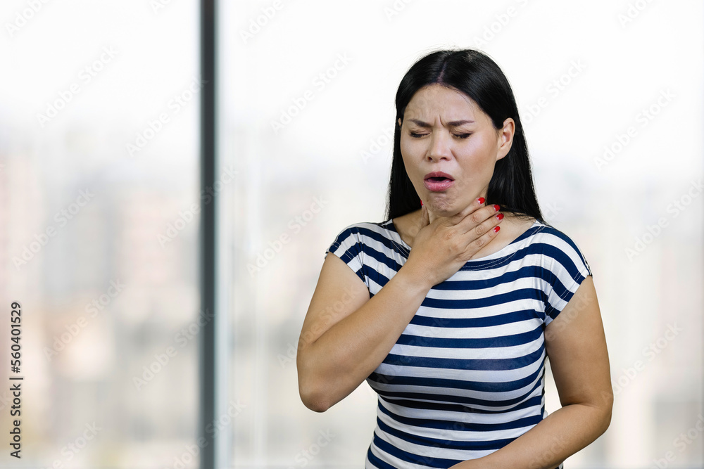 Young asian woman is coughing and touching her throat. Office windows background. Young asian woman is coughing and touching her throat. Office windows background.