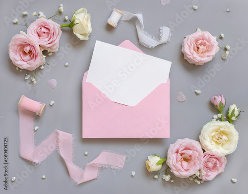 Blank card and envelope between pink roses and pink silk ribbons on grey top view, wedding mockup