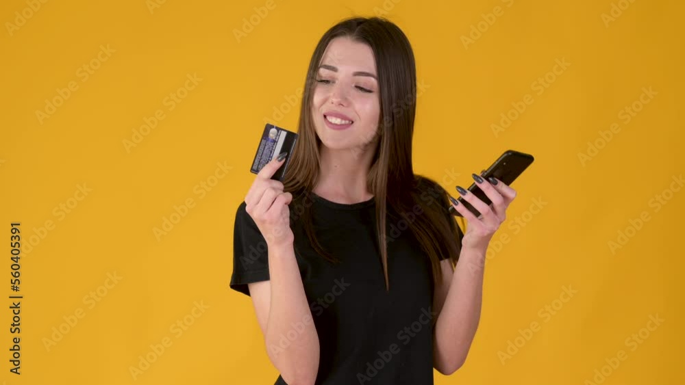 Cheerful young expressive girl Using mobile cell phone hold credit bank card makes purchases online shopping isolated on yellow background. People lifestyle concept. Cheerful young expressive girl Using mobile cell phone hold credit bank card makes purchases online shopping isolated on yellow background. People lifestyle concept.