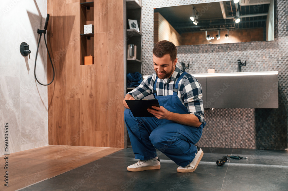 Sitting on the floor with notepad in hands. Plumber in blue uniform is at work in the bathroom
