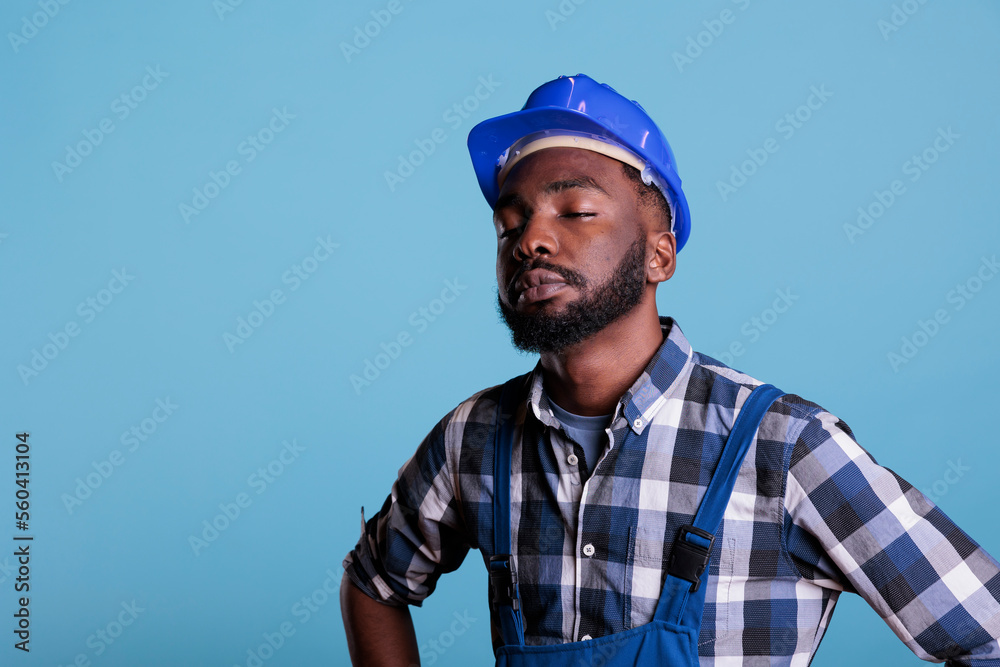 Fototapeta premium Tired exhausted construction worker in construction helmet and coveralls. African american employee with closed eyes feeling fatigued after hard work isolated on blue background.