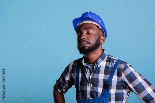 Tired exhausted construction worker in construction helmet and coveralls. African american employee with closed eyes feeling fatigued after hard work isolated on blue background. © DC Studio