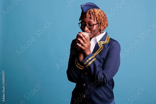 African american female flight attendant enjoying a cup of hot coffee before passengers board the plane. Stewardess wearing work uniform in studio shot against blue background.