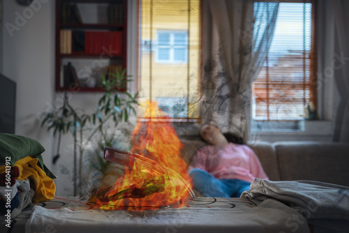 Hot flaming iron on ironing board. In defocused background woman is sleeping on sofa. Concept of fire hazard from electric household appliances