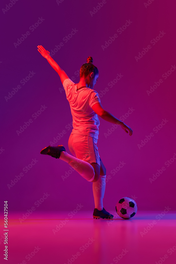 Young professional female football, soccer player in motion, training, playing over gradient pink background in neon light. Back view