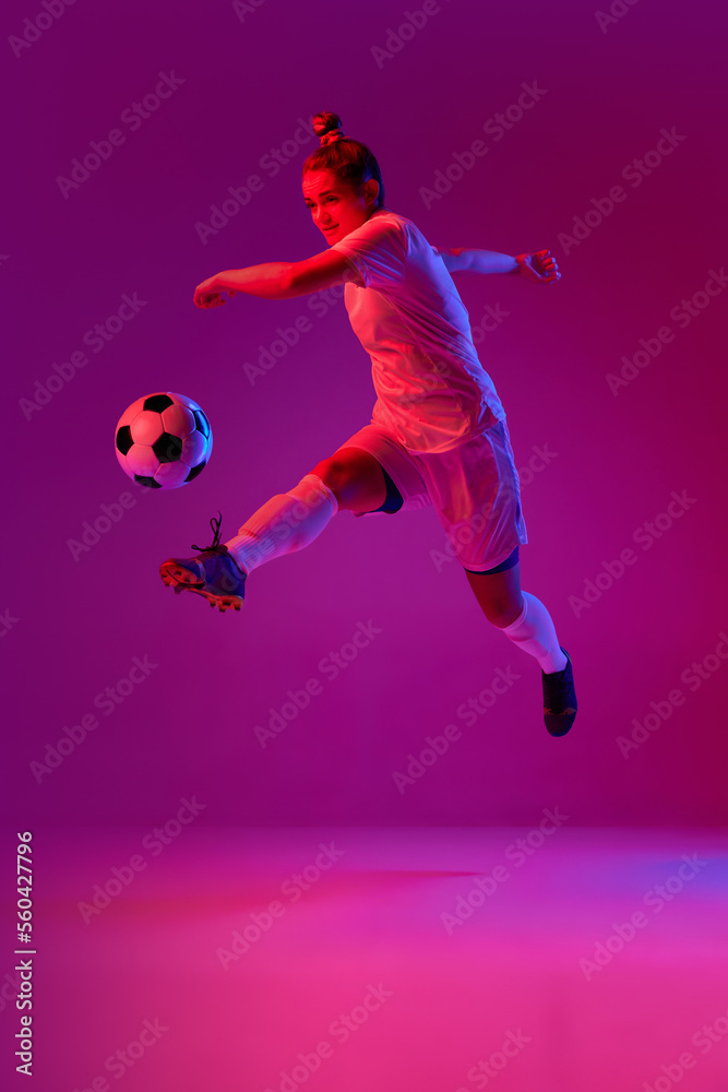 Young professional female football, soccer player in motion, training, playing over gradient pink background in neon light. Jump kick