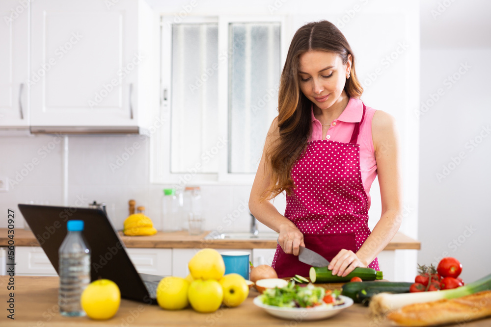 Young housewife using laptop and cooking in home kitchen