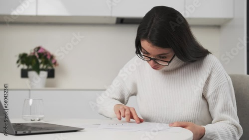 Young lady manager in black-rimmed glasses works at home checking data in papers. Focused woman sits at table near laptop in stylish kitchen working photo