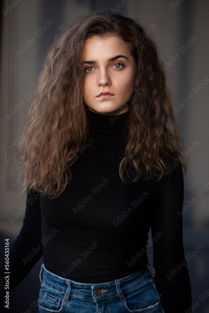 Naklejka premium Front photo of a feminity young woman with curly hair, wear black suitcase and jeans, looking at the side, posing near grey wall.