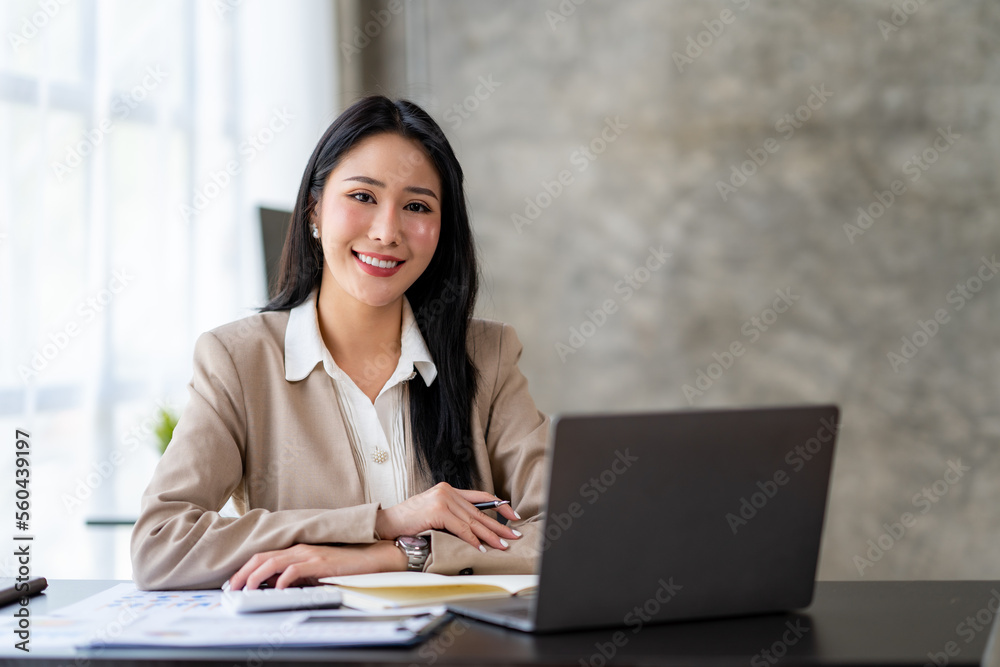 Asian young woman businesswoman using a laptop sitting smiling using modern analytical concepts While happily working at the desk in the office. Asian young woman businesswoman using a laptop sitting smiling using modern analytical concepts While happily working at the desk in the office.