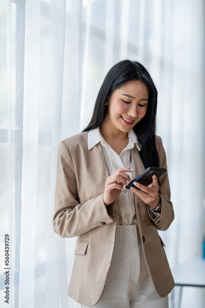 Obraz premium A beautiful Asian businesswoman stands and presses the phone to make customer contact explaining online marketing strategy, and finances in the office happily.