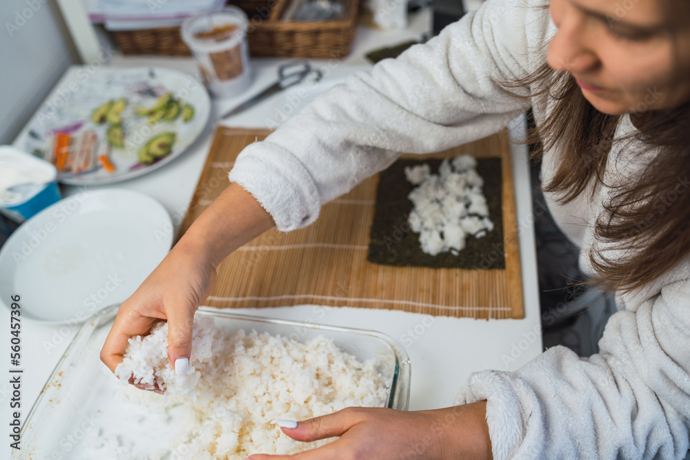 Woman taking a portion of rice from the pot to assemble a sheet of nori seaweed and prepare homemade sushi in her kitchen.