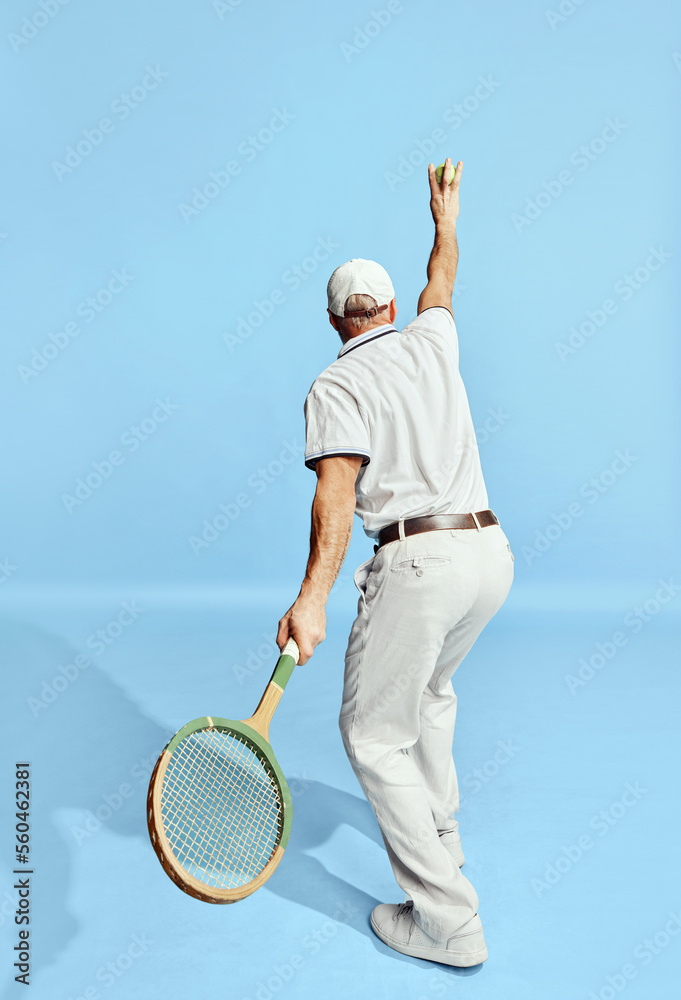 Back view. Serving. Portrait of handsome senior man in stylish white outfit playing tennis over blue background. Concept of leisure activity, hobby, lifestyle
