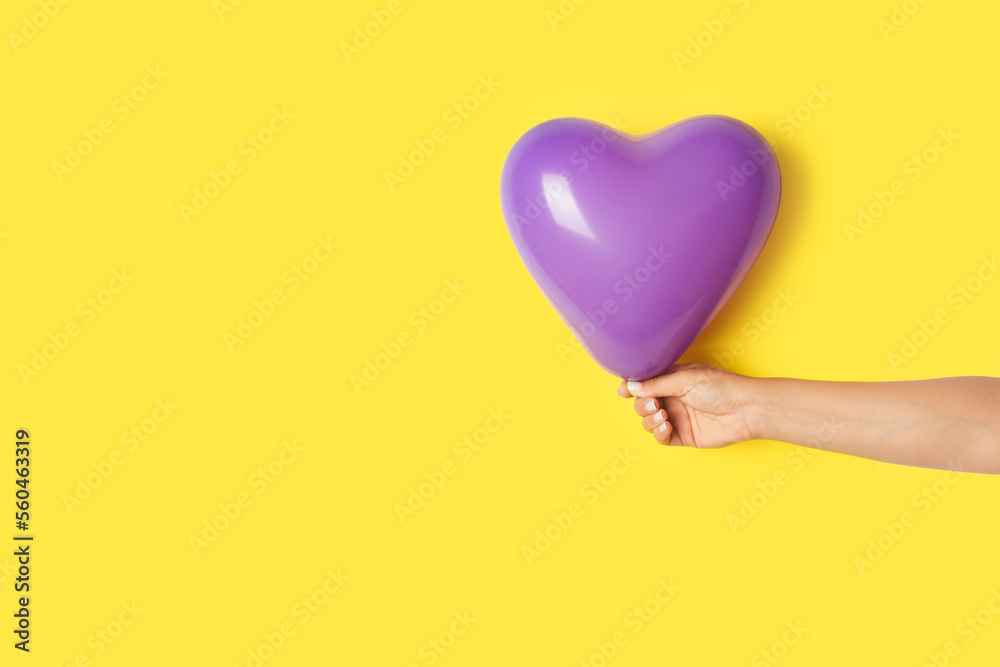 Fototapeta premium Mano de mujer sosteniendo un globo en forma de corazón de color morado sobre un fondo amarillo brillante liso y aislado. Vista de frente y de cerca. Copy space