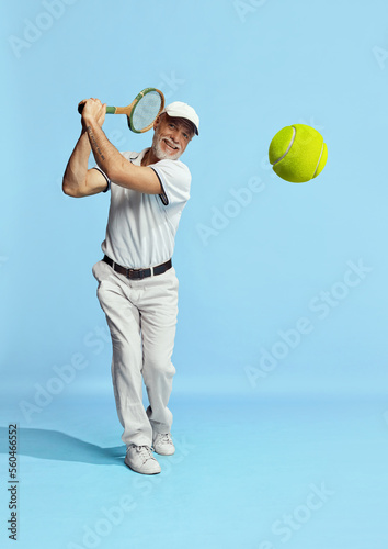 Tennis club member. Portrait of handsome senior man in stylish white outfit playing tennis over blue background. Concept of leisure activity, hobby, lifestyle © master1305