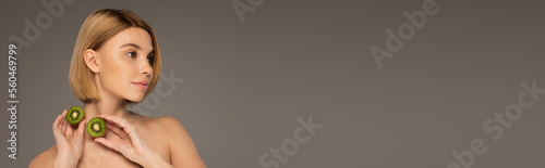 young woman with bare shoulders holding kiwi fruit and looking away isolated on grey, banner.