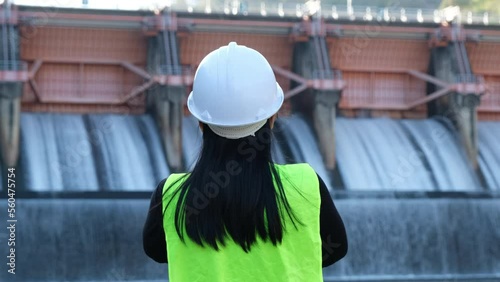 Rear view of female engineer in green vest and helmet standing outside against background of dam with hydroelectric power plant and irrigation.