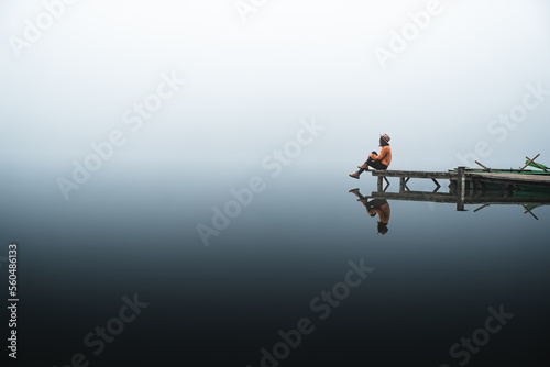 Side view of fashionable young woman standing on wooden dock looking at view on a misty morning. Female hipster relaxes on the edge of jetty admiring foggy landscape. Wonderful nature getaway