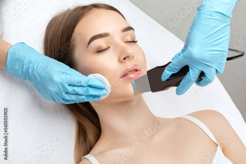 The doctor-cosmetologist makes the ultrasound cleaning procedure of the facial skin of a beautiful, young woman in a beauty salon. Cosmetology and professional skin care. 
