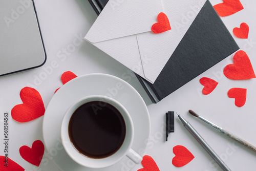 Home office desk with a cup of coffee, white envelope, laptop, red paper cut out hearts. Flat lay, love concept. Valentine's Day background.