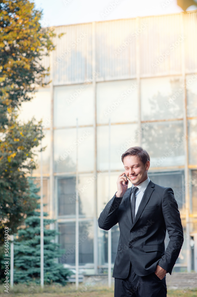 Happy young businessman in suit and tie talking on the mobile phone while standing outdoors with office building in the background