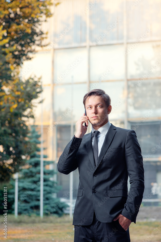 Confident young businessman in suit and tie talking on the mobile phone looking away while standing outdoors with office building in the background Confident young businessman in suit and tie talking on the mobile phone looking away while standing outdoors with office building in the background