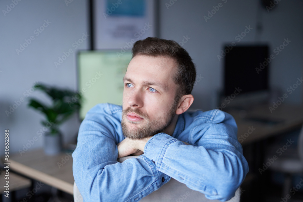 Cheerful bearded caucasian male sitting in home office looking at side dreaming. Young businessman or entrepreneur in denim shirt with computer monitor on background. High quality image Cheerful bearded caucasian male sitting in home office looking at side dreaming. Young businessman or entrepreneur in denim shirt with computer monitor on background. High quality image