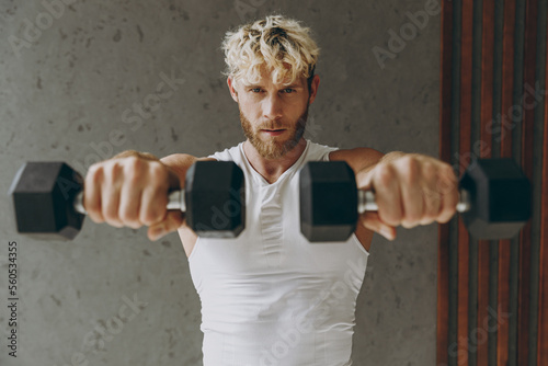 Confident calm caucasian young strong sporty athletic sportsman man wear white tank shirt lifting hold close up dumbbells stretch hands to camera warm up training indoor at gym. Workout sport concept. photo