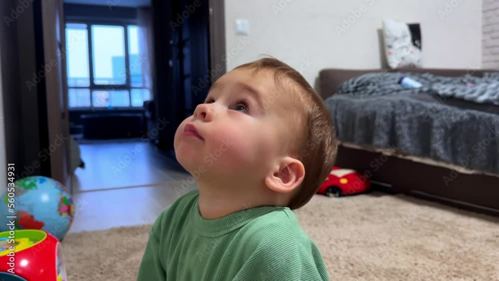 Little kid sits on the floor near his toys. Baby boy looks up with interest taking a finger into mouth. Close up side view. Little kid sits on the floor near his toys. Baby boy looks up with interest taking a finger into mouth. Close up side view.