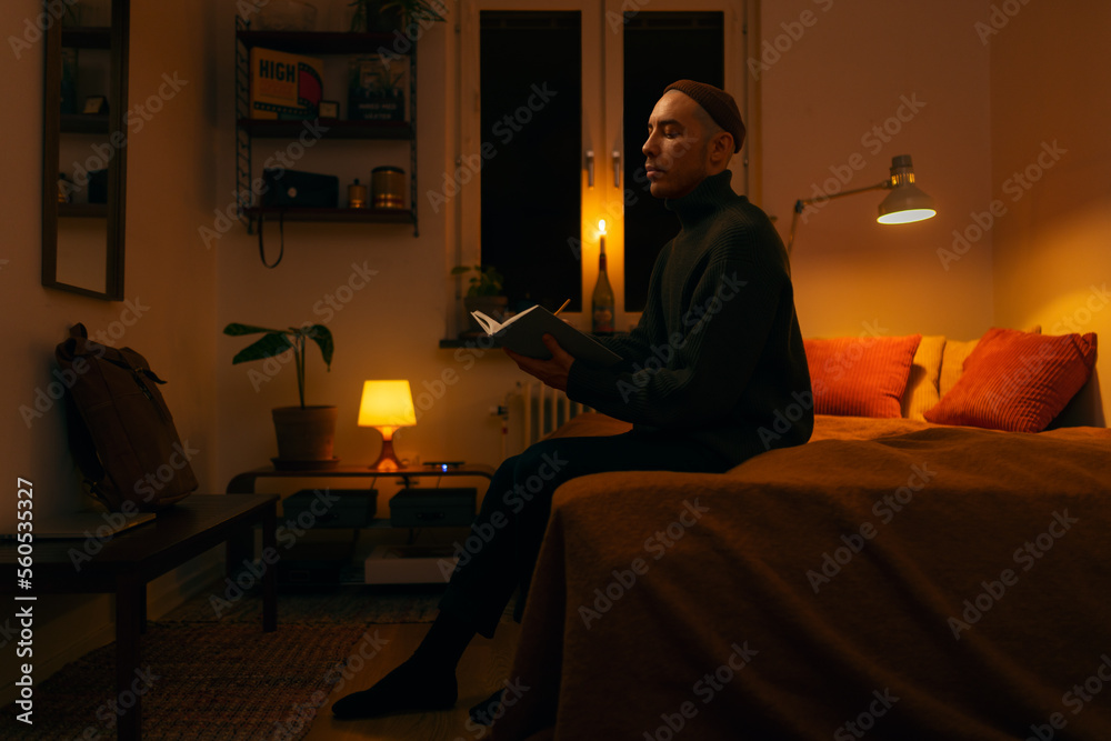 A caucasian man sitting on a bed in a bedroom writing in a book in the evening lit by a warm light.