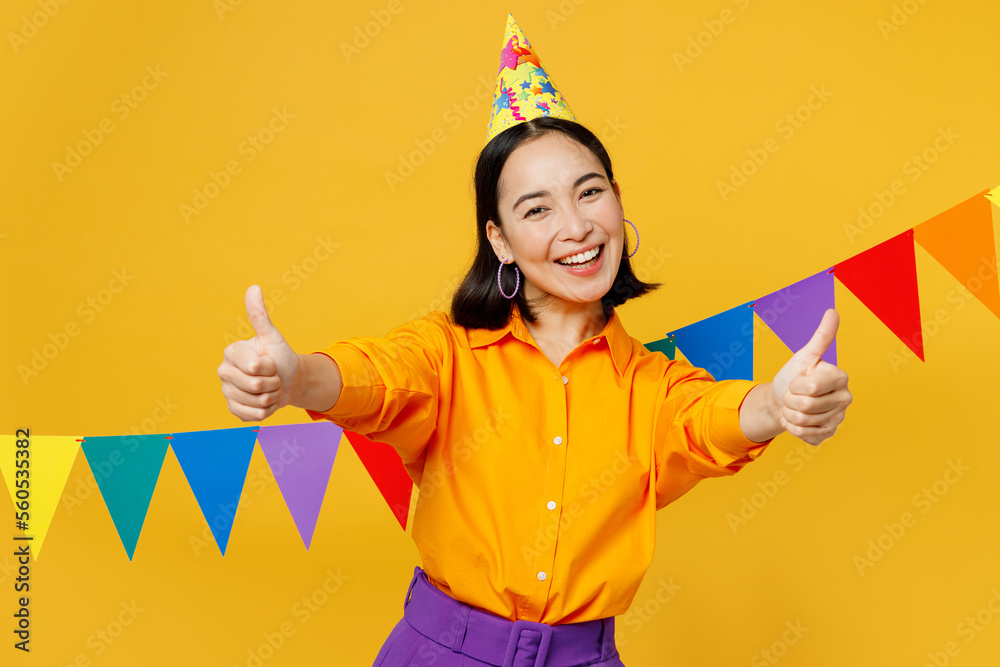 Happy fun satisfied young woman wears casual clothes hat celebrating near bunting flags show thumb up stretch hands to camera isolated on plain yellow background. Birthday 8 14 holiday party concept. Happy fun satisfied young woman wears casual clothes hat celebrating near bunting flags show thumb up stretch hands to camera isolated on plain yellow background. Birthday 8 14 holiday party concept.