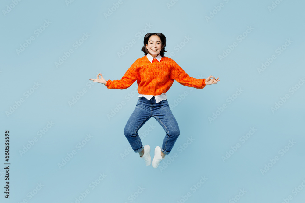 Full body spiritual young woman of Asian ethnicity wear orange sweater glasses jump high hold spreading hands in yoga om aum gesture relax meditate isolated on plain pastel light blue cyan background.