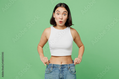 Young amazed surprised woman wear white clothes show loose pants on waist after weightloss isolated on plain pastel light green background. Proper nutrition healthy fast food unhealthy choice concept.