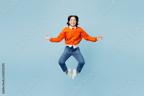 Full body spiritual young woman of Asian ethnicity wear orange sweater glasses jump high hold spreading hands in yoga om aum gesture relax meditate isolated on plain pastel light blue cyan background.